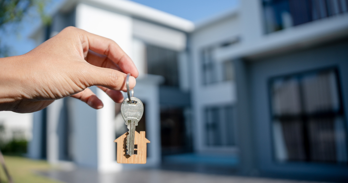 Foreground shows a hand holding a key with a wooden house keyring. The background is out of focus and is a new home.